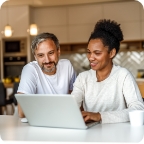 Two people sit in bright kitchen and use laptop.  